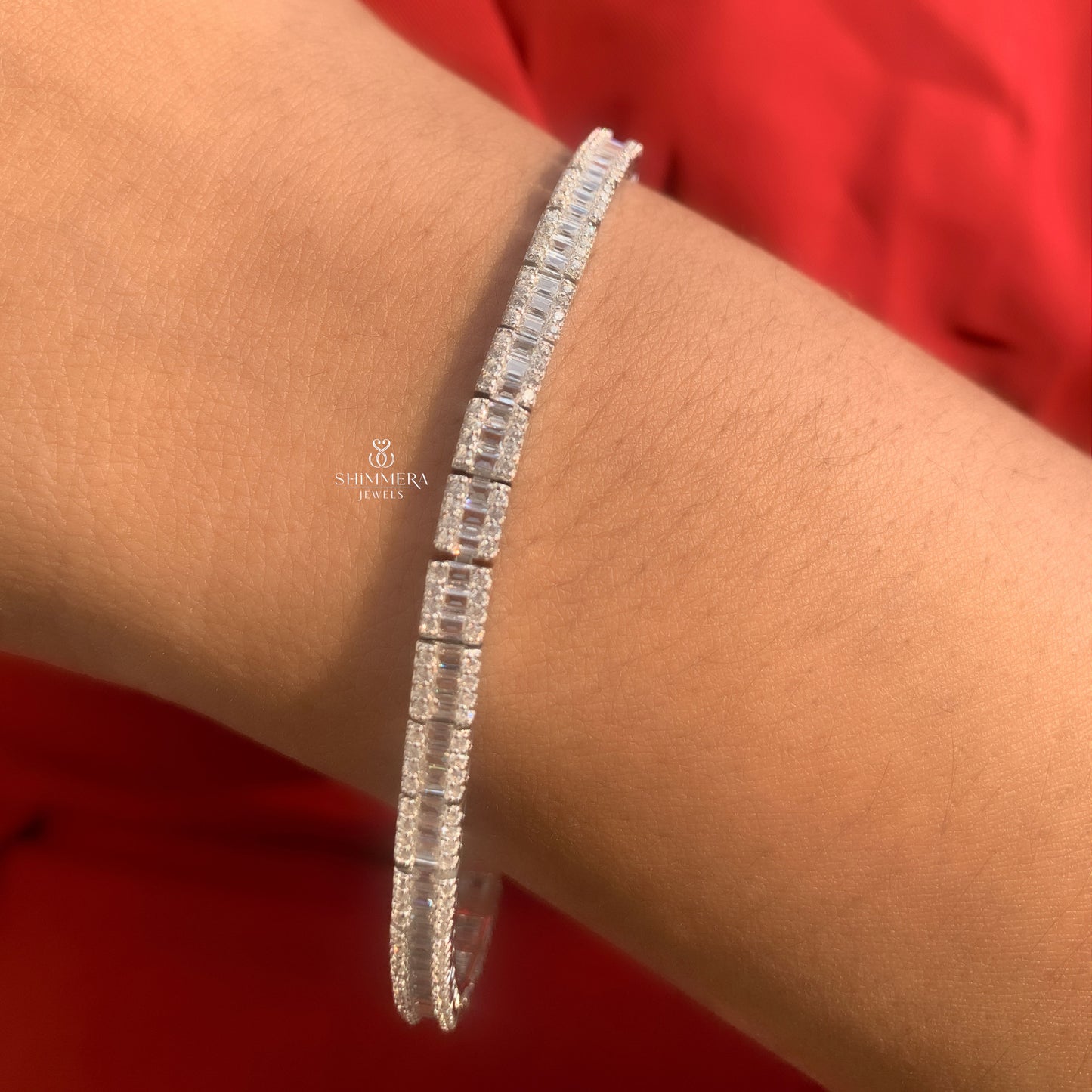Silver bracelet with clear stones on a wrist against a red background, featuring the Shri Kiran logo.