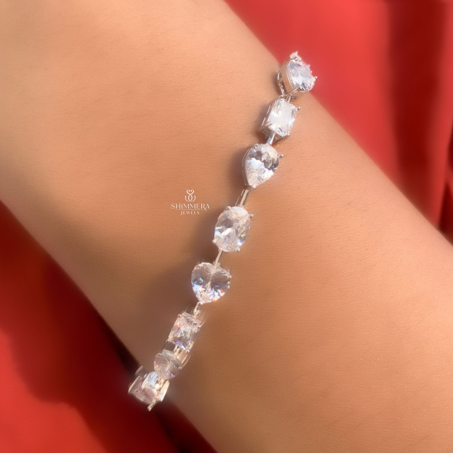 Silver bracelet with clear stones on a wrist against a red background, featuring the Shriharja Jewels logo.