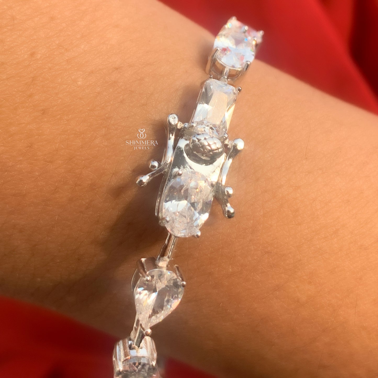 Silver bracelet with clear stones on a wrist against a red background, featuring the Shriekra Jewels logo.
