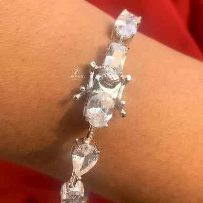Silver bracelet with clear stones on a wrist against a red background, featuring the Shriekra Jewels logo.